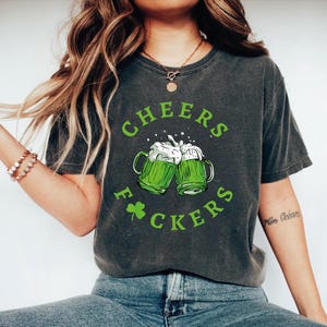 May include: Dark gray t-shirt with a green graphic. The graphic features two green beer mugs clinking together, with the words "CHEERS F*CKERS" in green, and a green shamrock. The shirt is a casual style.