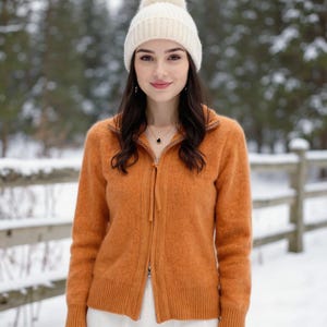 Puede incluir: Un suéter naranja con cremallera y un gorro de punto color crema con pompón. El suéter tiene un cuello alto y una cremallera en la parte delantera. El gorro es un cálido accesorio de invierno. El fondo es una escena nevada al aire libre.