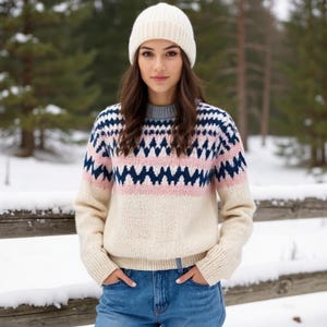 May include: A woman wearing a cream-coloured knitted beanie and a patterned jumper. The jumper features a geometric design in navy blue, pink, and white. She is also wearing blue jeans.