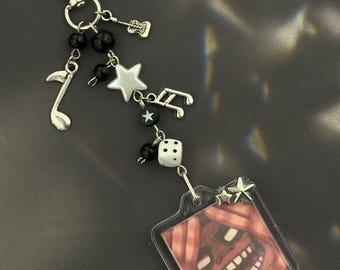 Puede incluir: Un llavero plateado con un mosquetón, cuentas negras y dijes de notas musicales. Cuenta con una etiqueta de acrílico transparente con una ilustración de cara de dibujos animados y detalles de estrellas, además de un dado blanco.