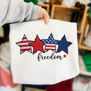 May include: A white sweatshirt featuring an embroidered design of four stars in a row. The stars alternate between red and white stripes and solid red or blue colours. The word freedom is embroidered in black cursive script below the stars.