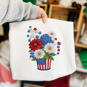 May include: White sweatshirt featuring a floral arrangement in a patriotic theme. The bouquet includes red dahlias, white daisies, blue hydrangeas, and small red and blue flowers, all arranged in a vase decorated with the American flag.