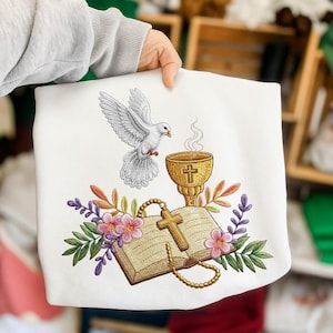 Puede incluir: Sudadera blanca con un diseño religioso bordado. El diseño incluye una paloma blanca, un cáliz dorado, un libro abierto con una cruz y detalles florales en rosa, morado y verde. La sudadera es sostenida por una persona.
