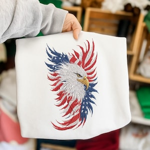 Op de afbeelding: Witte sweatshirt met een geborduurd adelaarsontwerp. De kop en veren van de adelaar zijn gekleurd in rood, wit en blauw, wat lijkt op de Amerikaanse vlag. De adelaar heeft een gele snavel op een witte achtergrond.