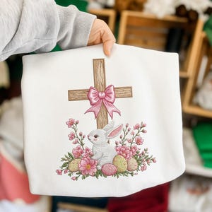 Może przedstawiać: Biała bluza z haftowanym wielkanocnym wzorem. Wzór zawiera drewniany krzyż z różową kokardą, białego królika, różowe kwiaty i zdobione pisanki. Bluza jest trzymana przez osobę.