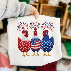 Può includere: Felpa bianca con un ricamo di tre galli in rosso, bianco e blu, con fuochi d'artificio sopra. I galli sono decorati con stelle e strisce, che rappresentano la bandiera americana. Il design è patriottico e festoso.