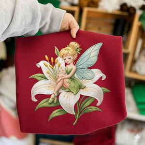 May include: A red fabric item featuring a detailed illustration of a fairy sitting on a white lily flower. The fairy has blonde hair, green clothing, and blue wings. The lily has yellow stamens and green leaves.