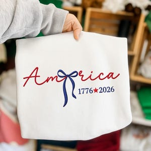 May include: A white sweatshirt featuring the word "America" embroidered in red script, a blue bow, and the numbers "1776" and "2026" with a red star. The sweatshirt is held by a person.