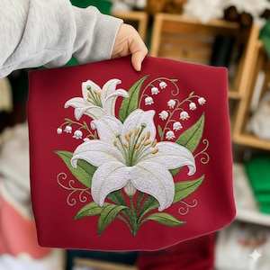 May include: A red textile featuring an embroidered floral design. The embroidery showcases white lilies and lily of the valley, with green leaves and stems. The design is detailed with yellow stamens and delicate swirls.