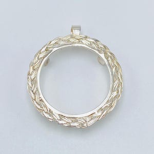 Op de afbeelding: Een zilverkleurige hanger met een cirkelvormig gevlochten ontwerp. De hanger heeft een kleine lus aan de bovenkant voor een ketting. Het gevlochten patroon creëert een textuur, en het ontwerp is eenvoudig en elegant.