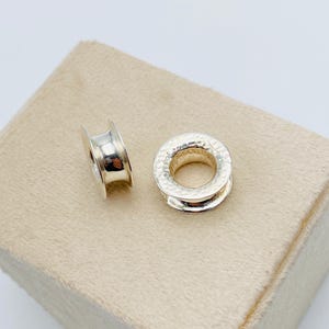 May include: Two silver-colored metal eyelets with a cylindrical shape and a central opening. One eyelet is positioned upright showing a smooth interior, while the other lies flat, displaying a textured, hammered finish on its circular top surface.