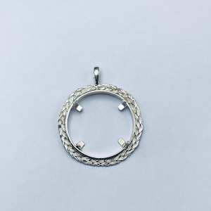 May include: A silver pendant with a circular design. The pendant has a braided rope-like border. Four small, square prongs are inside the circle. A small loop is at the top for a chain. The pendant is designed to hold a circular object.