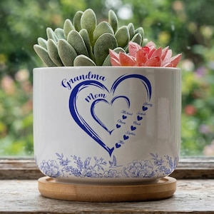 May include: White ceramic planter with a blue heart design and the words "Grandma" and "Mom". The planter includes names and is filled with succulents. It sits on a wooden coaster.