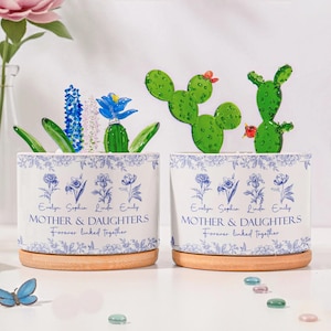 May include: Two white ceramic planters with blue floral designs and wooden bases. Each planter has the text "MOTHER & DAUGHTERS" and "Forever linked together." One planter has glass cactus and flower accents.