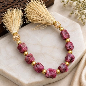 Peut inclure: Un cordon décoratif composé de perles de rubis brut rose foncé, alternées avec de petites perles sphériques dorées lisses et des perles filigranées dorées texturées, terminé aux deux extrémités par de longs pompons métalliques dorés.