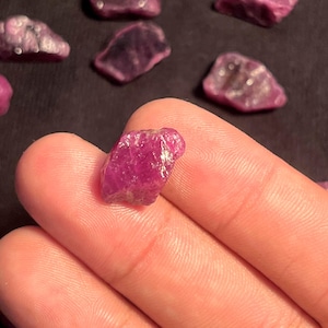 Peut inclure: Une petite pierre précieuse brute d'un rouge rosé profond avec une texture de surface irrégulière et facettée. La pierre est tenue sur un fond sombre, avec plusieurs autres pierres roses brutes similaires éparpillées en arrière-plan.