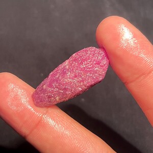 May include: A raw, unpolished ruby crystal with a vibrant magenta-pink hue and a textured, crystalline surface. The stone has an elongated, irregular shape with visible natural facets and mineral inclusions throughout its structure.
