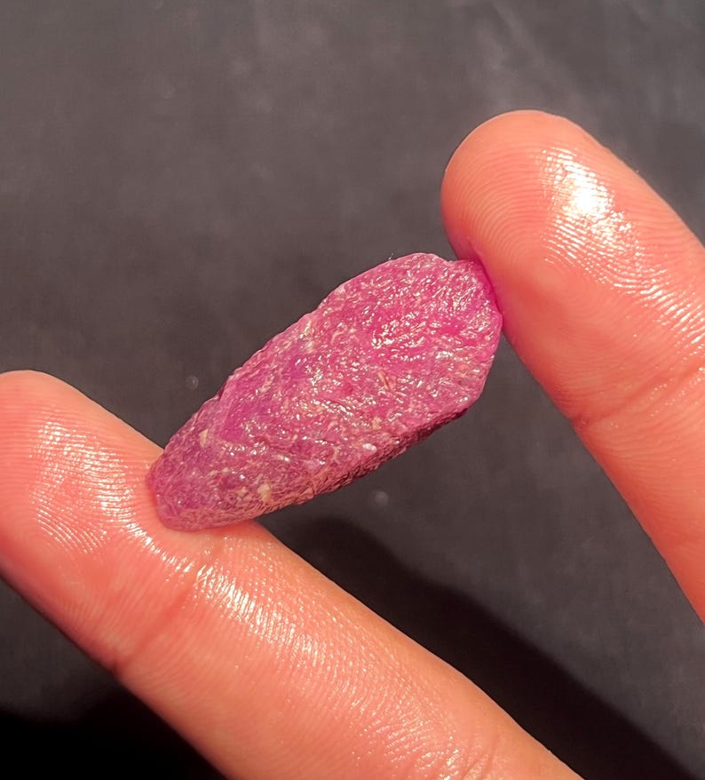 May include: A raw, unpolished ruby crystal with a vibrant magenta-pink hue and a textured, crystalline surface. The stone has an elongated, irregular shape with visible natural facets and mineral inclusions throughout its structure.