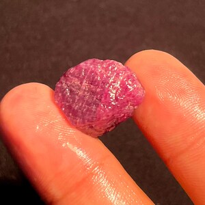 May include: A raw, unpolished ruby gemstone with a deep magenta-pink hue and a textured, crystalline surface. The stone displays natural facets and irregular edges, reflecting light across its translucent, vibrant, and saturated mineral structure.