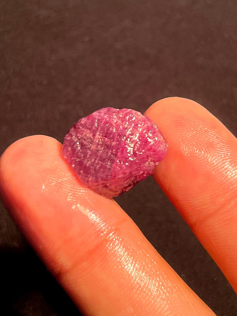 May include: A raw, unpolished ruby gemstone with a deep magenta-pink hue and a textured, crystalline surface. The stone displays natural facets and irregular edges, reflecting light across its translucent, vibrant, and saturated mineral structure.