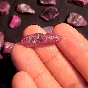 Op de afbeelding: Een kleine, langwerpige, ruwe robijnkristal met een diepe magenta-rode tint en een getextureerd, kristallijn oppervlak. De steen is scherp in beeld gebracht en toont natuurlijke facetten en onregelmatige randen van een ongeslepen edelsteen.