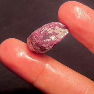 Peut inclure: Une pierre précieuse brute et non polie aux teintes magenta et violet profond. La pierre présente une surface irrégulière et texturée avec des facettes naturelles et des structures cristallines qui captent la lumière, révélant des nuances de rose foncé.