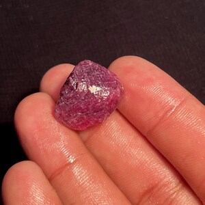 May include: A raw, unpolished gemstone with a deep magenta and reddish-purple hue. The stone features a rough, textured surface with natural facets and a small, reflective white inclusion on its upper face, showcasing its organic, uncut form.
