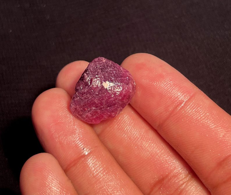 May include: A raw, unpolished gemstone with a deep magenta and reddish-purple hue. The stone features a rough, textured surface with natural facets and a small, reflective white inclusion on its upper face, showcasing its organic, uncut form.