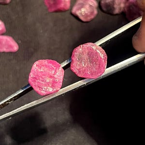 K&ouml;nnte beinhalten: Zwei rohe, leuchtend pink-rote Rubine, gehalten von einer silberfarbenen Metallpinzette. Die Edelsteine haben eine unregelm&auml;&szlig;ige, abgerundete Form mit einer texturierten, kristallinen Oberfl&auml;che. Mehrere &auml;hnliche lose Rubine liegen verstreut.