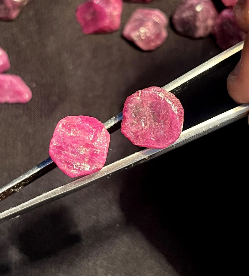 K&ouml;nnte beinhalten: Zwei rohe, leuchtend pink-rote Rubine, gehalten von einer silberfarbenen Metallpinzette. Die Edelsteine haben eine unregelm&auml;&szlig;ige, abgerundete Form mit einer texturierten, kristallinen Oberfl&auml;che. Mehrere &auml;hnliche lose Rubine liegen verstreut.