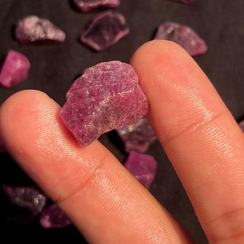 May include: A raw, unpolished ruby gemstone with a deep magenta-pink hue and a textured, crystalline surface. The stone displays natural facets and irregular edges, showcasing a translucent quality when light hits the mineral structure.
