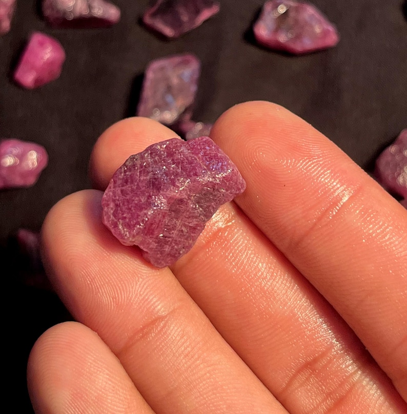 May include: A raw, unpolished ruby gemstone with a deep magenta-red hue and a textured, crystalline surface. The stone features natural facets and irregular edges, showcasing a translucent quality when light hits the mineral structure.