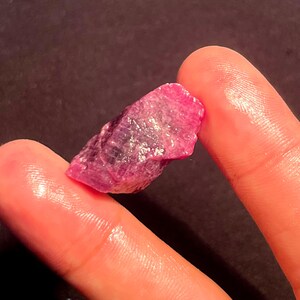 May include: A raw, unpolished ruby gemstone with a vibrant magenta-pink hue and a jagged, crystalline texture. The stone features natural facets and translucent areas that catch the light, displaying deep red and pink color variations throughout.
