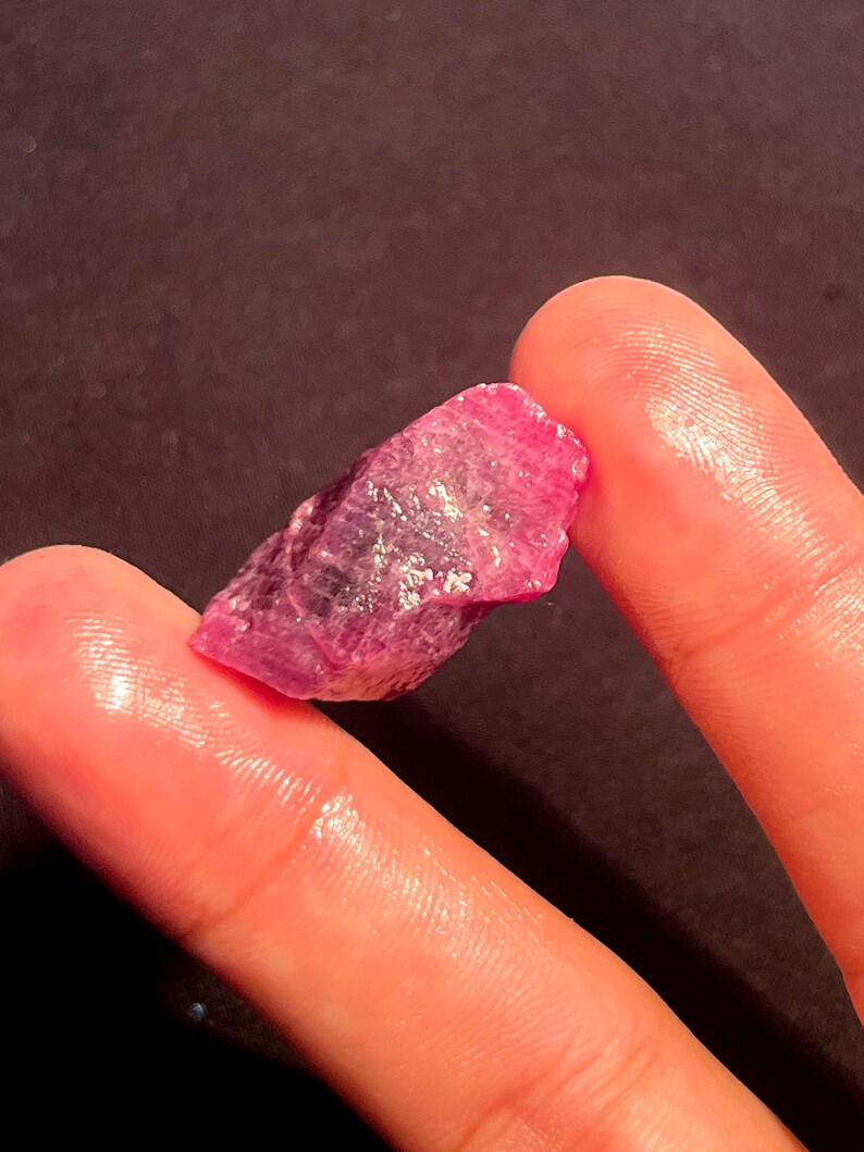 May include: A raw, unpolished ruby gemstone with a vibrant magenta-pink hue and a jagged, crystalline texture. The stone features natural facets and translucent areas that catch the light, displaying deep red and pink color variations throughout.