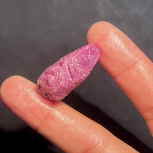 May include: A raw, unpolished ruby crystal with a vibrant magenta-pink hue and a textured, crystalline surface. The stone has an elongated, irregular shape with natural facets and a semi-translucent appearance, showcasing deep pink colour variations.