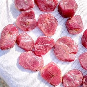 Könnte beinhalten: Eine Sammlung roher, ungeschliffener Rubin-Edelsteine in verschiedenen Formen und Größen. Die Edelsteine zeigen einen tiefen, rötlich-rosa Farbton und liegen auf einer weißen Stoffoberfläche. Die raue Textur und die natürlichen Formen der Rubine sind deutlich sichtbar.