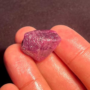 May include: A raw, unpolished ruby gemstone with a deep magenta and purple hue. The stone features a natural, irregular crystalline structure with visible facets and a semi-translucent appearance, showcasing a rough, textured surface texture.