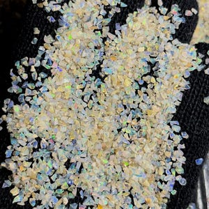 Könnte beinhalten: Nahaufnahme eines Haufens kleiner, schillernder Opalchips. Die Chips sind eine Mischung aus Weiß, Creme und Hellblau, mit Regenbogenfarben. Die Chips sind auf einer schwarzen Oberfläche verstreut.