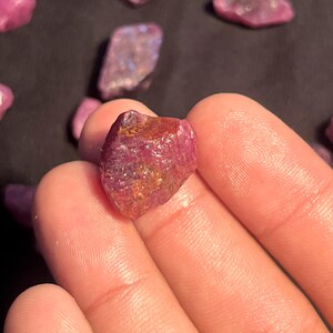 K&ouml;nnte beinhalten: Ein roher, unpolierter Rubin-Edelstein mit einem tiefen Magenta- und Karmesin-Farbton, der eine texturierte, kristalline Oberfl&auml;che mit nat&uuml;rlichen Facetten und Einschl&uuml;ssen aufweist und seine unregelm&auml;&szlig;ige, kantige Form sowie seine Transluzenz zeigt.