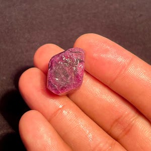May include: A raw, unpolished gemstone with a deep magenta and pink hue. The stone features a multifaceted, irregular surface with visible crystalline textures and natural inclusions, showcasing a translucent quality throughout its structure.