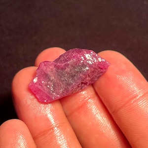 May include: A raw, unpolished ruby gemstone with a deep pinkish-red hue and a textured, crystalline surface. The stone features irregular edges and natural facets that catch the light, displaying variations in color intensity and clarity.