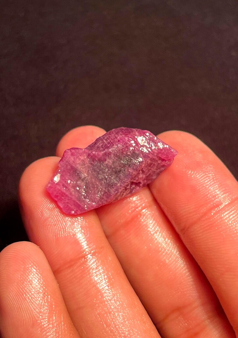 May include: A raw, unpolished ruby gemstone with a deep pinkish-red hue and a textured, crystalline surface. The stone features irregular edges and natural facets that catch the light, displaying variations in color intensity and clarity.