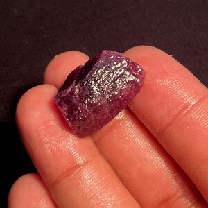 May include: A raw, unpolished ruby gemstone with a deep magenta and purple hue. The stone features a rough, crystalline texture with natural facets and an irregular shape, displaying a translucent quality that catches light across its surface.