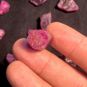 Könnte beinhalten: Ein kleiner, roher, tief pink-roter Edelstein mit einer texturierten, kristallinen Oberfläche. Der Stein hat eine unregelmäßige, abgerundete Form mit sichtbaren Facetten und inneren Lichtreflexionen, die eine lebendige, gesättigte Farbe zeigen.