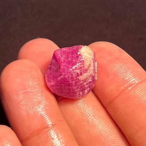 Peut inclure: Une pierre précieuse brute et non taillée d'une teinte magenta et rose profond vibrante. La pierre présente une forme irrégulière à facettes avec des textures cristallines visibles et une petite tache d'inclusion minérale blanche opaque.