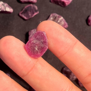 Puede incluir: Una pequeña piedra preciosa en bruto de color rojo rosado intenso con una forma irregular y multifacética. La piedra tiene una calidad translúcida con texturas internas e inclusiones naturales visibles, mostrando un color vibrante y saturado en toda su superficie.