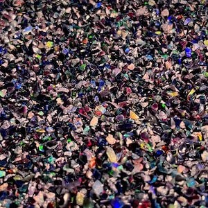 May include: A collection of small, crushed black opal fragments featuring a dark base color with iridescent flashes of blue, green, red, and yellow. The irregular, jagged pieces are densely packed together, creating a textured surface.