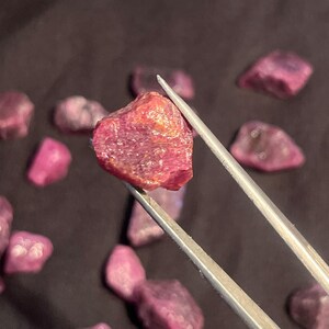 K&ouml;nnte beinhalten: Ein roher, tief pink-roter Edelstein, der mit einer Metallpinzette gehalten wird. Der Stein hat eine raue, kristalline Textur mit nat&uuml;rlichen Facetten und internen Lichtreflexionen. Mehrere &auml;hnliche, unpolierte rote Mineralfragmente liegen verstreut.