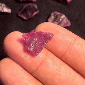 Peut inclure: Un petit fragment de pierre précieuse brut, d'un rouge rosé profond, avec une texture cristalline dentelée et des bords translucides. La pierre présente des facettes naturelles et des reflets internes, révélant une couleur vive et saturée.