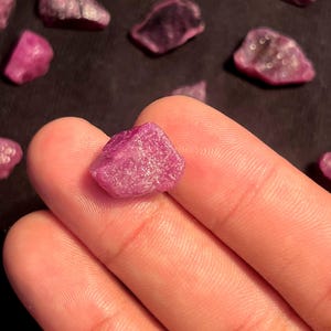 May include: A small, raw, unpolished ruby gemstone with a vibrant magenta-pink hue and a textured, crystalline surface. The stone features irregular, jagged edges and a natural, opaque appearance, showcasing the raw mineral structure of the crystal.